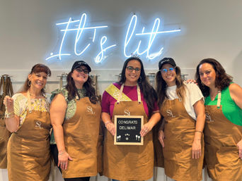 Five smiling women wearing brown aprons pose inside a craft studio under a blue neon sign reading It's lit, holding a small letter board with a congratulatory message — group workshop celebration photo.