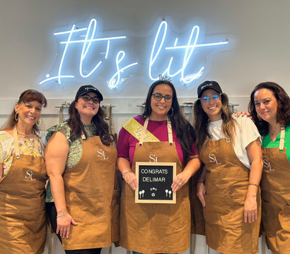 Five smiling women wearing brown aprons pose inside a craft studio under a blue neon sign reading It's lit, holding a small letter board with a congratulatory message — group workshop celebration photo.