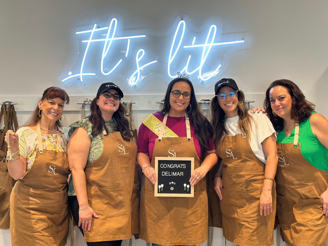 Five smiling women wearing brown aprons pose inside a craft studio under a blue neon sign reading It's lit, holding a small letter board with a congratulatory message — group workshop celebration photo.