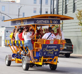 Group of women celebrating a bachelorette party on a yellow pedal bar party bike in a downtown street, pedaling and raising drinks while wearing casual outfits and cowboy hats.