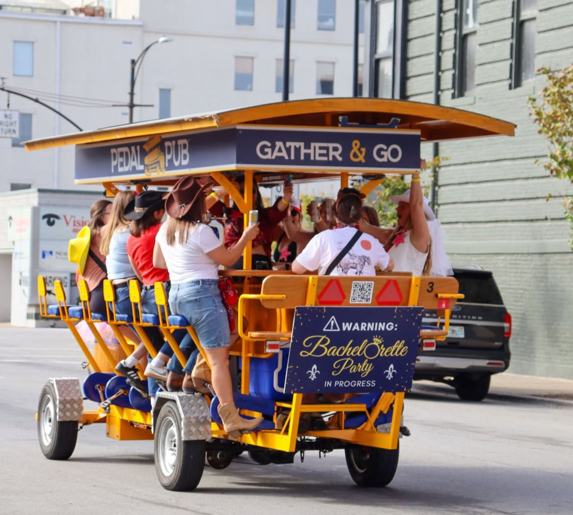 Group of women celebrating a bachelorette party on a yellow pedal bar party bike in a downtown street, pedaling and raising drinks while wearing casual outfits and cowboy hats.