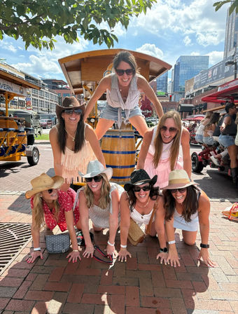 Seven women in summer outfits and cowgirl hats laughing as they form a playful human pyramid on a sunny downtown brick plaza, with pedal-tour vehicles and city buildings in the background.