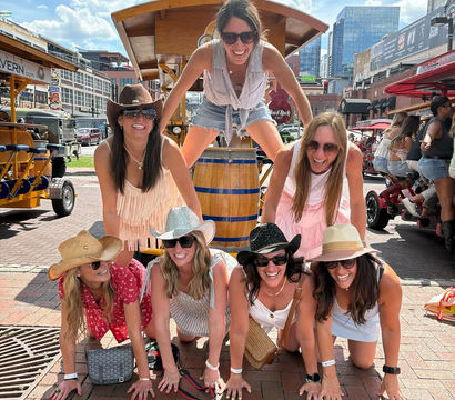 Seven women in summer outfits and cowgirl hats laughing as they form a playful human pyramid on a sunny downtown brick plaza, with pedal-tour vehicles and city buildings in the background.
