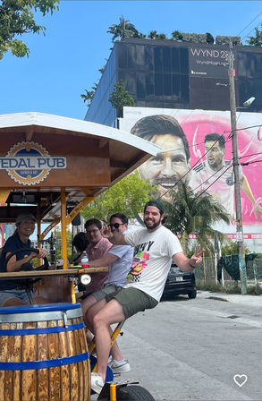 Cheerful group riding a pedal-powered bar on a sunny urban street with palm trees and a large pink mural of a soccer player on a building wall.