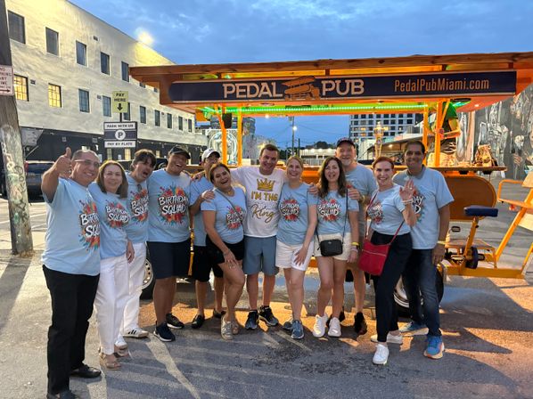 Smiling birthday group in matching “Birthday Squad” shirts posing in front of a brightly lit orange pedal-powered party bar on a Miami street at dusk.