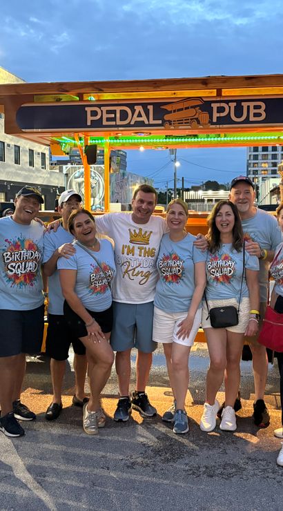 Smiling birthday group in matching “Birthday Squad” shirts posing in front of a brightly lit orange pedal-powered party bar on a Miami street at dusk.