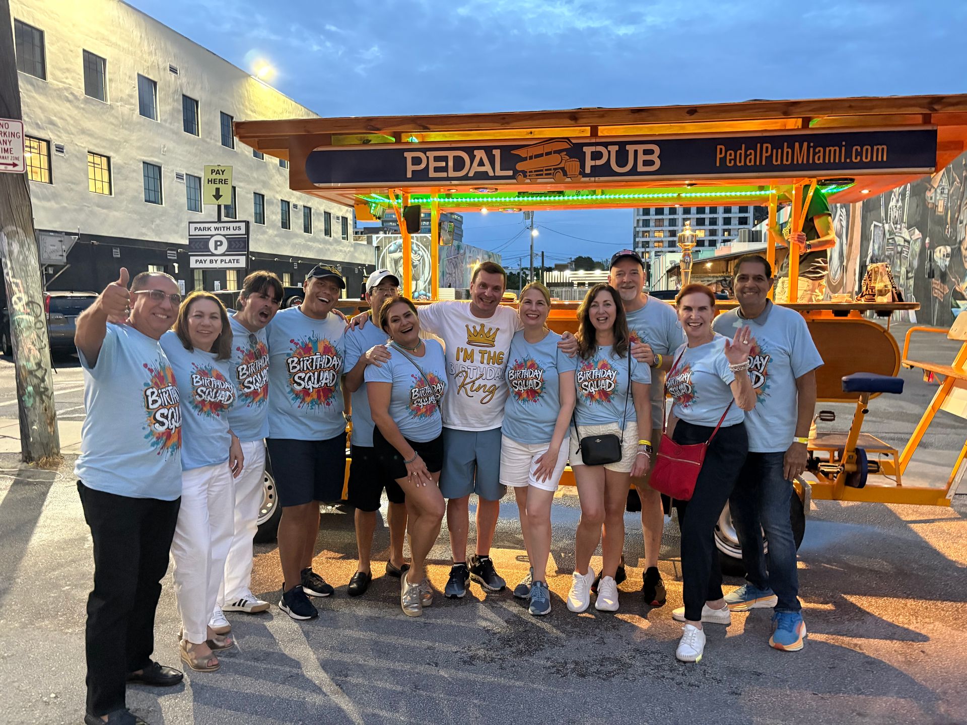 Smiling birthday group in matching “Birthday Squad” shirts posing in front of a brightly lit orange pedal-powered party bar on a Miami street at dusk.