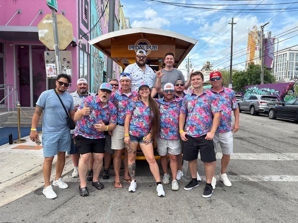 Group of people in matching bright tropical shirts posing in front of a wooden pedal-powered party bike on a sunny urban street lined with colorful murals and parked cars.