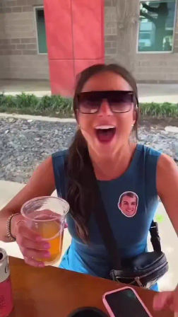 Laughing woman in sunglasses on an outdoor patio holding a plastic cup of orange drink, wearing a blue sleeveless top with a novelty face sticker, smartphone and fanny pack on the table, modern brick building in the background.