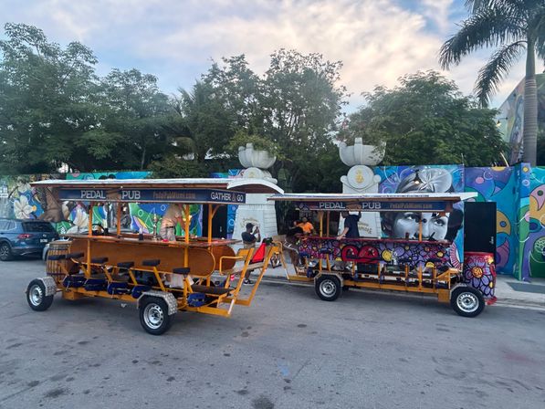 Two colorful pedal-powered party bikes parked on a mural-lined tropical urban street under palm trees at dusk, riders socializing at the built-in bar seats.