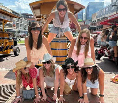 Seven women in summer outfits and cowboy hats forming a playful human pyramid on a brick sidewalk in a sunny downtown entertainment district with bars and skyline buildings