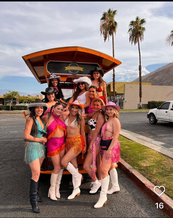 Group of women in colorful cowgirl outfits, fringe skirts and boots posing on and around an orange pedal-party vehicle in a sunny desert town with palm trees and mountains in the background.