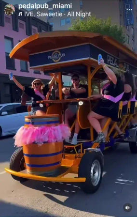 Group of people riding an orange pedal-powered party bike decorated with a pink tutu and sashes, cheering and holding drinks on a sunny Miami street.