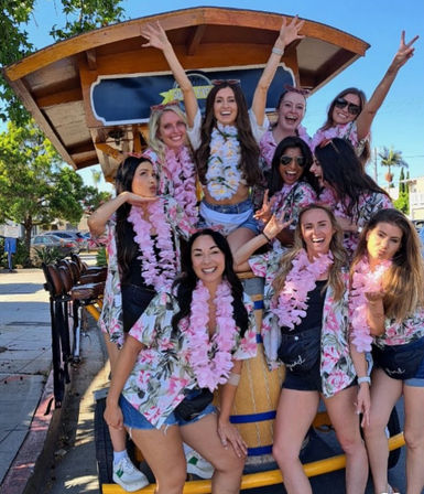 Group of women in floral shirts and pink leis laughing and cheering on a pedal-powered party cart on a sunny street, friends celebrating outdoors.