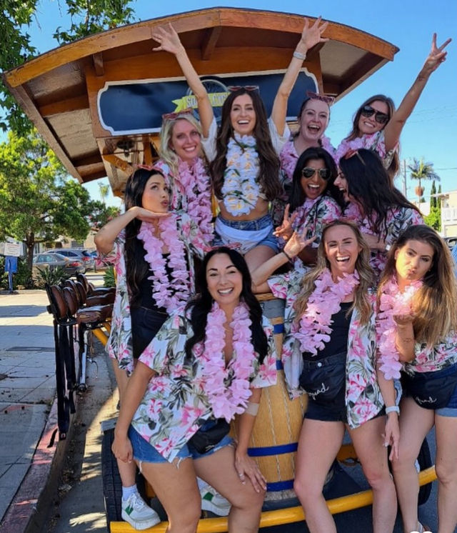 Group of women in floral shirts and pink leis laughing and cheering on a pedal-powered party cart on a sunny street, friends celebrating outdoors.