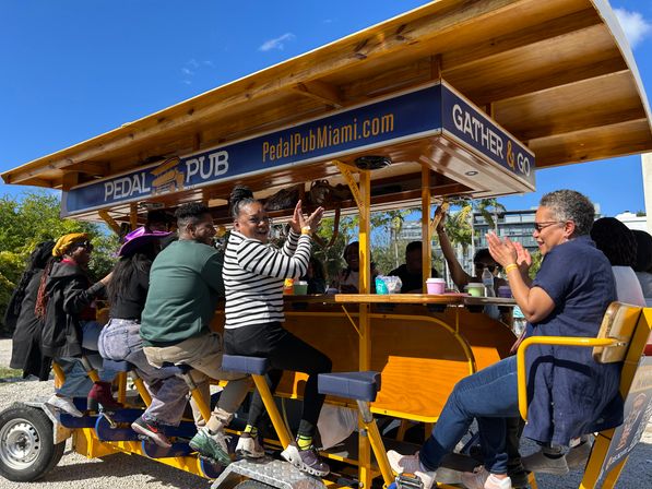 Group of friends laughing and clapping on a yellow pedal-powered party pub bike with palm trees and a bright blue sky in Miami, outdoor social ride scene