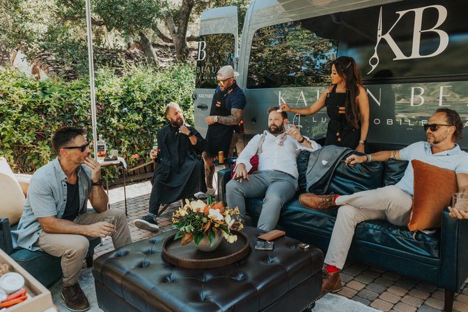 Sunlit backyard patio lounge with five men relaxing on leather sofas, sipping drinks while a mobile grooming team trims beards beside a branded salon van and a flower-topped ottoman.