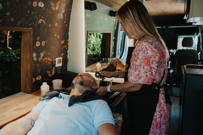Stylist in a floral dress washing a client's hair inside a converted mobile salon van, with butterfly wallpaper, mirror, sink and grooming bottles visible.