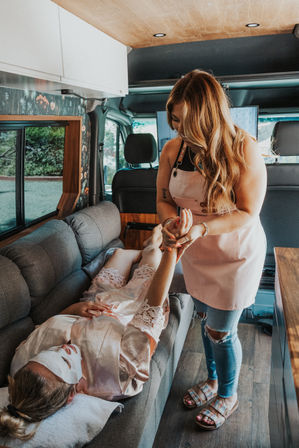 Client wearing a sheet facial mask and silk robe reclines on a couch inside a cozy campervan while a technician in a pink apron gives a relaxing hand massage — mobile spa van pampering and wellness on the go.