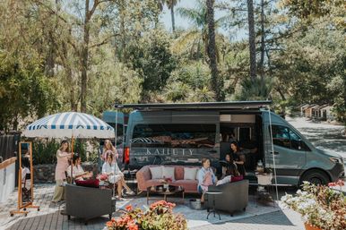 Mobile luxury salon van on a sunny patio with palm trees; women getting hair and makeup under a striped umbrella and lounging on a pink sofa.