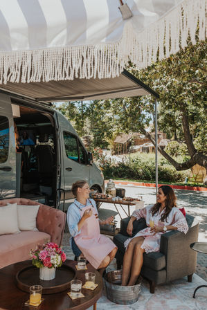 Two women relaxing under a striped fringe awning beside a camper van at a sunny outdoor campsite — one gives a foot soak while they chat over drinks, snacks, and flowers.