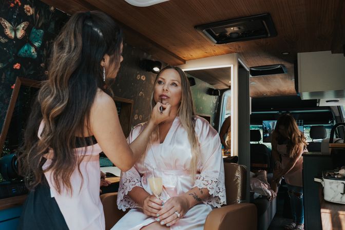 Woman in pink satin robe holding a champagne flute while a makeup artist applies lipstick inside a stylish mobile beauty van during wedding prep