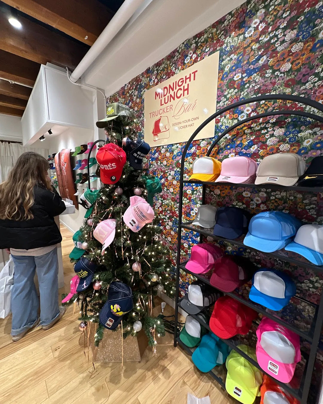 Colorful trucker hats displayed on a decorated holiday tree and neatly stacked on shelves in a floral-walled boutique with a design-your-own trucker hat sign and a shopper browsing.