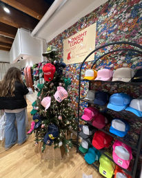 Colorful trucker hats displayed on a decorated holiday tree and neatly stacked on shelves in a floral-walled boutique with a design-your-own trucker hat sign and a shopper browsing.