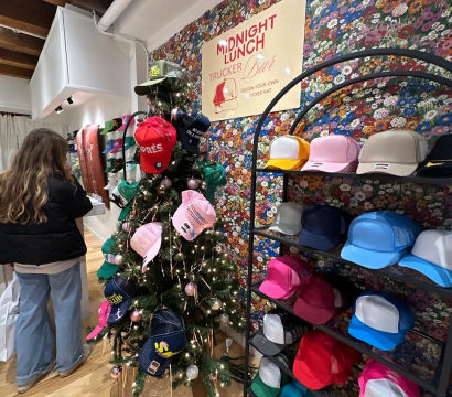 Colorful trucker hats displayed on a decorated holiday tree and neatly stacked on shelves in a floral-walled boutique with a design-your-own trucker hat sign and a shopper browsing.