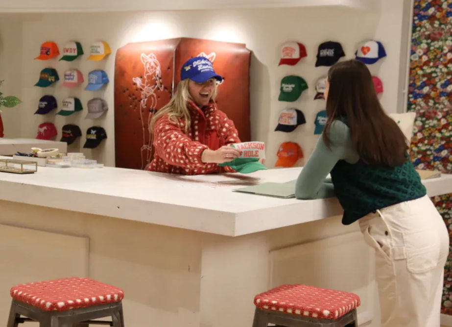 Bright hat shop interior with a smiling clerk handing a 'Jackson Hole' cap to a leaning customer across a white counter, colorful caps on the wall and red-cushioned stools in the foreground.