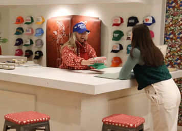 Bright hat shop interior with a smiling clerk handing a 'Jackson Hole' cap to a leaning customer across a white counter, colorful caps on the wall and red-cushioned stools in the foreground.