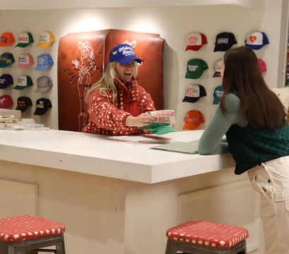 Bright hat shop interior with a smiling clerk handing a 'Jackson Hole' cap to a leaning customer across a white counter, colorful caps on the wall and red-cushioned stools in the foreground.