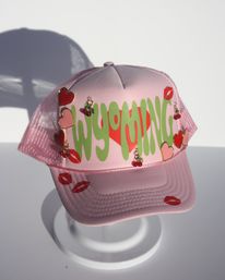 Pink mesh trucker hat with bold green 'WYOMING' lettering, red heart and cherry charms, and red kiss-print stickers displayed on a white stand.