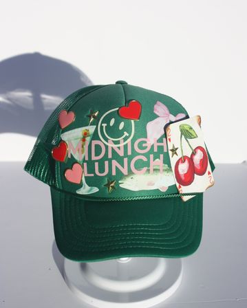 Green mesh trucker hat on white stand with pink 'MIDNIGHT LUNCH' text, smiley-face print, martini and fish graphics, heart pins and star studs, and a 2 of hearts playing card featuring cherries attached — playful product shot.