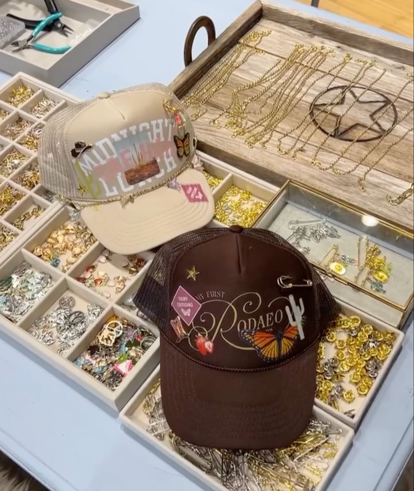 Two boho western trucker hats with butterfly and cactus patches displayed on a boutique jewelry tray filled with gold chains, charms, and pins.
