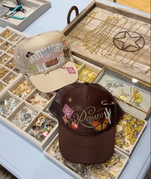 Two boho western trucker hats with butterfly and cactus patches displayed on a boutique jewelry tray filled with gold chains, charms, and pins.