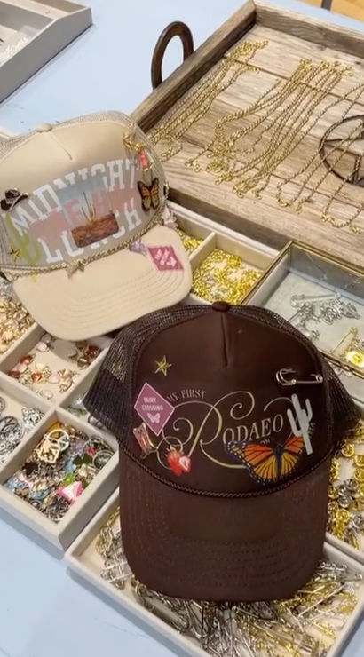 Two boho western trucker hats with butterfly and cactus patches displayed on a boutique jewelry tray filled with gold chains, charms, and pins.