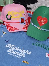 Two playful trucker hats — pink cap with yellow script and red leaf embroidery and green cap with a red heart — displayed on a denim shirt printed with New York City motifs, a yellow taxi and cocktail graphic.