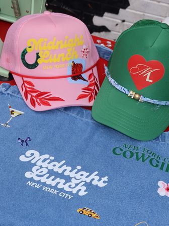 Two playful trucker hats — pink cap with yellow script and red leaf embroidery and green cap with a red heart — displayed on a denim shirt printed with New York City motifs, a yellow taxi and cocktail graphic.