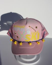 Pink mesh trucker hat with APRES SKI graphic, gold star studs and safety-pin accent, blue beaded cord strung with tiny yellow rubber duck charms, photographed on a white surface with soft shadow.