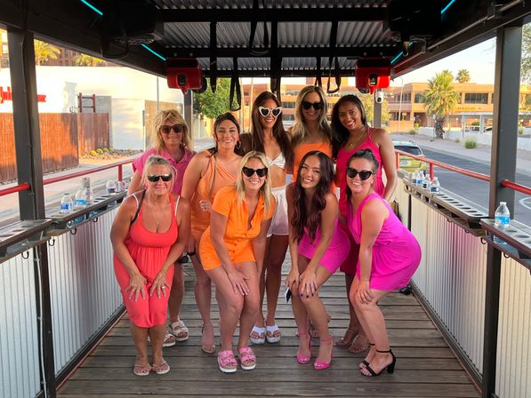 Nine women in bright summer dresses and sunglasses posing and smiling on a covered party trolley/pedal pub along a sunlit downtown street with palm trees and water bottles on the rail — lively friends' outing.