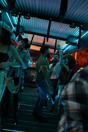 Neon-lit nightlife scene with people dancing and laughing under blue and orange lights in a covered outdoor bar area, some wearing cowboy hats and casual denim.