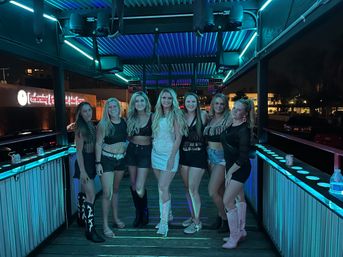 Seven women posing on a neon‑lit rooftop patio at night, wearing party outfits and cowboy boots, center woman in a white dress and sparkly boots under teal and purple LED lights