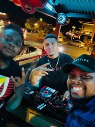 Three friends smiling around a DJ controller on an outdoor patio at night, urban streetlights and cars in the background