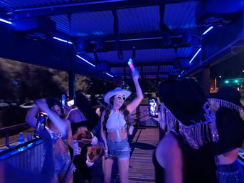 Neon-lit outdoor deck party at night with friends dancing; woman in a white cowboy hat and heart-shaped sunglasses waves a light-up stick while others film and hold novelty props.
