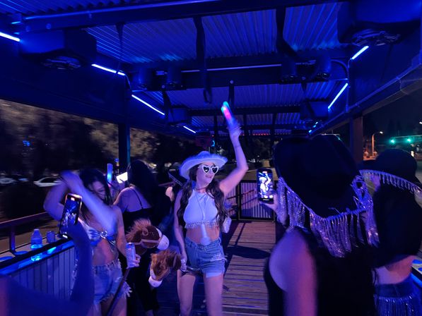 Neon-lit outdoor deck party at night with friends dancing; woman in a white cowboy hat and heart-shaped sunglasses waves a light-up stick while others film and hold novelty props.