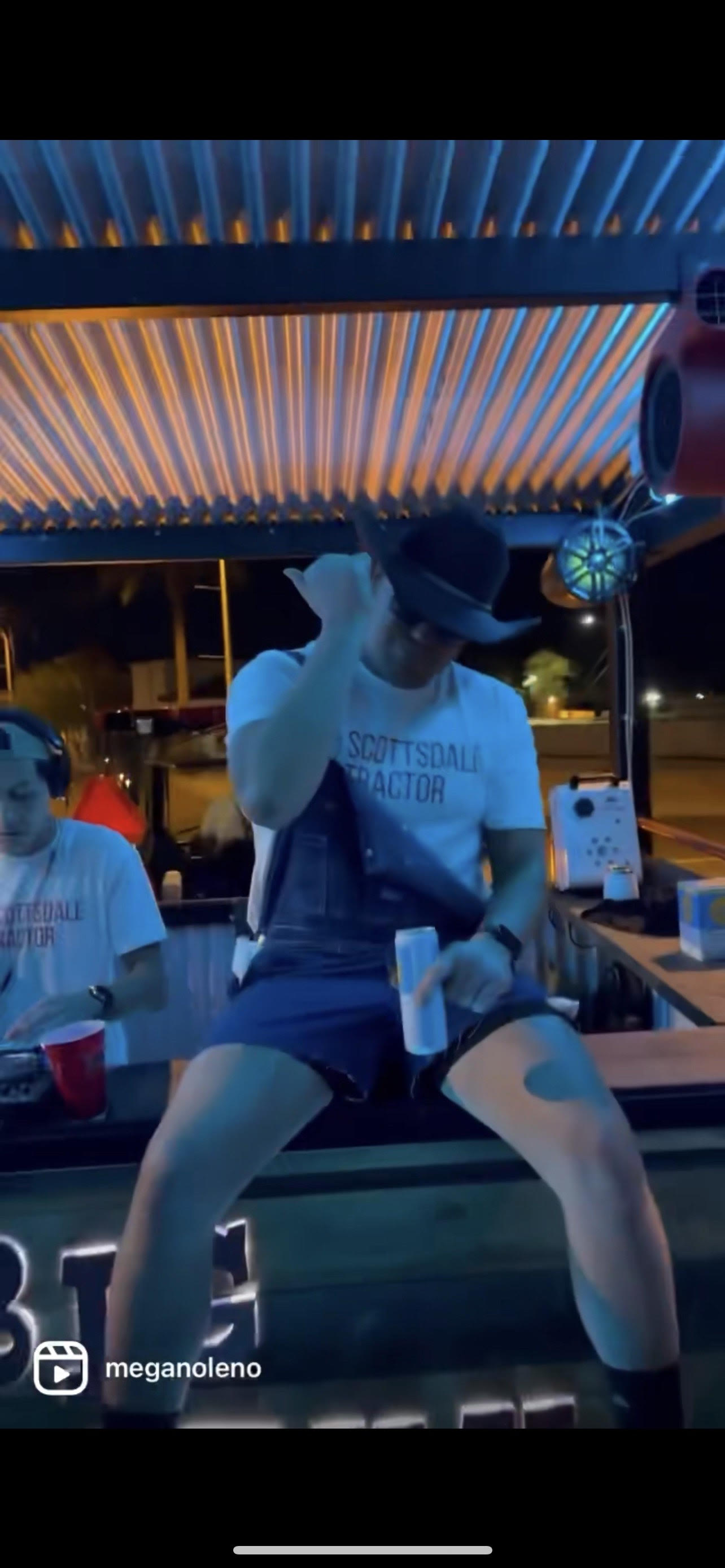 Nighttime outdoor bar patio in Scottsdale-style setting with blue-orange corrugated roof lights, a man in a cowboy hat perched on the counter holding a canned drink while a DJ mixes to his left.
