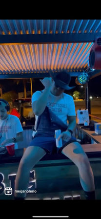 Nighttime outdoor bar patio in Scottsdale-style setting with blue-orange corrugated roof lights, a man in a cowboy hat perched on the counter holding a canned drink while a DJ mixes to his left.