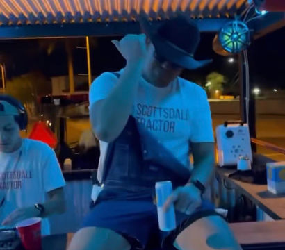 Nighttime outdoor bar patio in Scottsdale-style setting with blue-orange corrugated roof lights, a man in a cowboy hat perched on the counter holding a canned drink while a DJ mixes to his left.