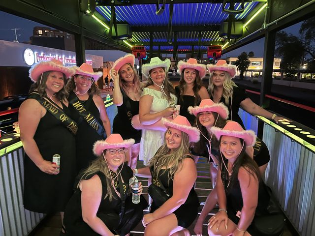 Bachelorette party on a lit outdoor bar patio at night — a smiling group of women in black dresses and pink fuzzy cowboy hats with the bride in white and sashes, posing for a fun group photo